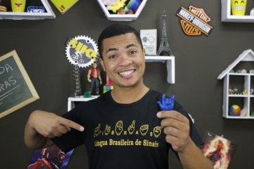 Pessoa sorrindo ensinando Libras, com sinais de mãos claros e fundo vibrante, representando o curso Libras Intensive de Rodrigo Timóteo.