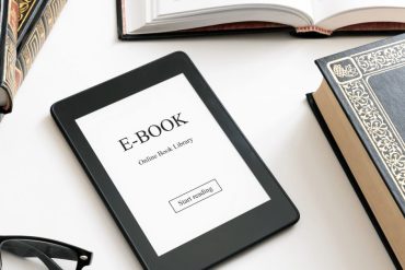 Homem sorrindo, segurando um e-book e apontando para uma tela de computador com gráficos de vendas, representando o sucesso em vender e-books online.